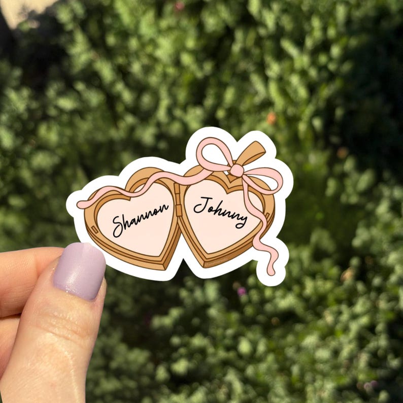 May include: A sticker featuring two heart-shaped lockets, each with a name inside. The lockets are light brown with pink interiors and a pink ribbon bow. The names "Shannon" and "Johnny" are written in cursive.