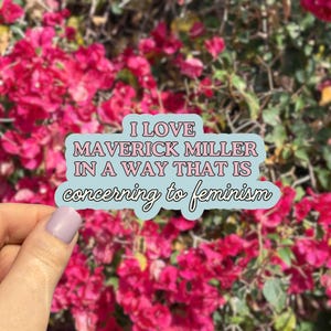 May include: A sticker with the text "I LOVE MAVERICK MILLER IN A WAY THAT IS concerning to feminism" in pink and white on a light blue background. The sticker is held in front of a background of pink flowers.