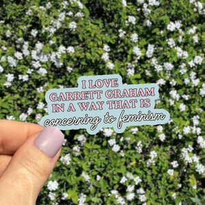 May include: A light blue sticker with the text "I LOVE GARRETT GRAHAM IN A WAY THAT IS concerning to feminism" in pink and white. The sticker is held in front of a background of green foliage with small white flowers.