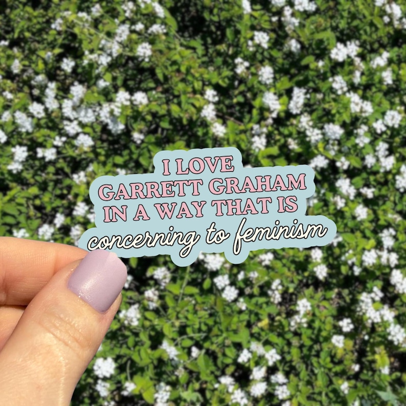 May include: A light blue sticker with the text "I LOVE GARRETT GRAHAM IN A WAY THAT IS concerning to feminism" in pink and white. The sticker is held in front of a background of green foliage with small white flowers.
