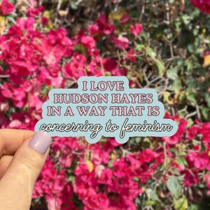May include: A sticker with the text "I LOVE HUDSON HAYES IN A WAY THAT IS concerning to feminism" in pink and black lettering on a light blue background. The sticker is held in front of a background of bright pink flowers.