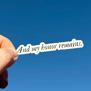 May include: A white sticker with the text "And my honor remains." is held against a bright blue sky. The sticker has a white border and the text is in a simple, elegant font.