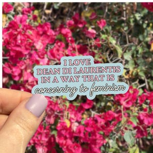 Puede incluir: Una pegatina con el texto "I LOVE DEAN DI LAURENTIS IN A WAY THAT IS concerning to feminism" en rosa y blanco sobre un fondo azul claro. La pegatina se sostiene frente a un fondo de flores rosas.