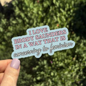 May include: A light blue sticker with the text "I LOVE BRODY SAUNDERS IN A WAY THAT IS concerning to feminism" in pink and white. The sticker is held by a hand with a light purple nail polish.