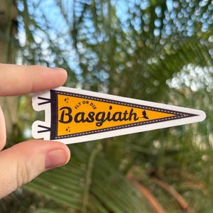 May include: A yellow and white pennant-shaped sticker with the text "Basgiath" in a bold, black font. The sticker also includes the phrase "Fly or Die" in smaller black text. The background is a blurred view of green foliage.
