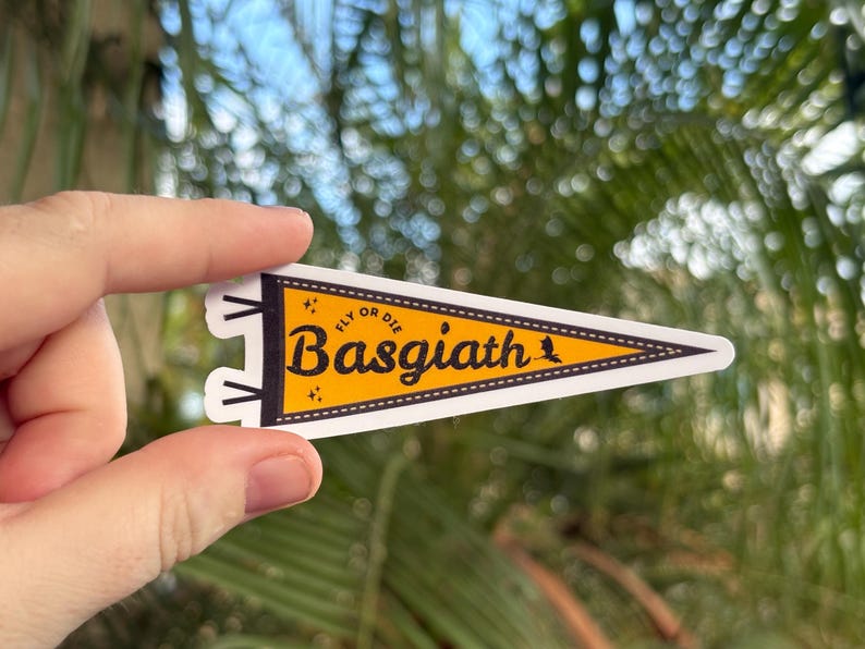 May include: A yellow and white pennant-shaped sticker with the text "Basgiath" in a bold, black font. The sticker also includes the phrase "Fly or Die" in smaller black text. The background is a blurred view of green foliage.