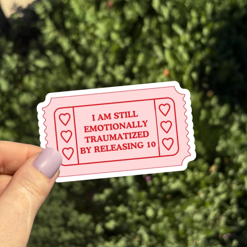 May include: A pink ticket-shaped sticker with the text "I AM STILL EMOTIONALLY TRAUMATIZED BY RELEASING 10" in red. The sticker has a scalloped edge and three heart shapes on each side.