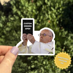Pu&ograve; includere: Un adesivo con l'immagine di Papa Francesco che tiene in mano un cartello nero con la scritta "CUSTOM ORDERS OPEN". L'adesivo &egrave; tenuto da una mano con lo smalto viola chiaro. &Egrave; visibile anche una grafica a forma di stella gialla con la scritta "QUESTIONS? MESSAGE ME!"