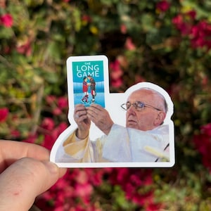 May include: A sticker featuring an image of the Pope holding up a sticker that says "The Long Game". The sticker within the sticker depicts two hockey players. The background is blurred with pink flowers and green foliage.