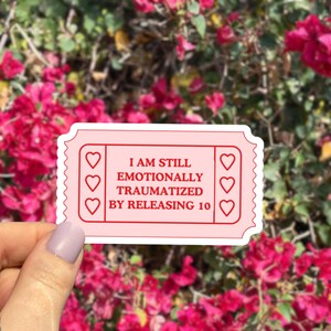 May include: A pink sticker in the shape of a ticket with the text "I AM STILL EMOTIONALLY TRAUMATIZED BY RELEASING 10". The sticker has three heart shapes on each side.