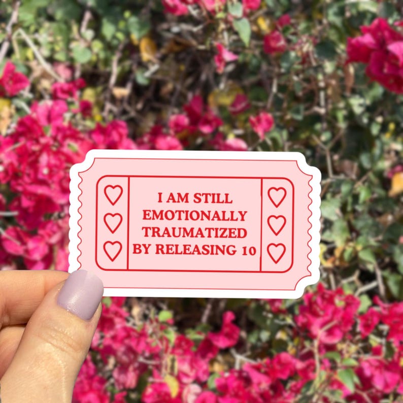 May include: A pink sticker in the shape of a ticket with the text "I AM STILL EMOTIONALLY TRAUMATIZED BY RELEASING 10". The sticker has three heart shapes on each side.