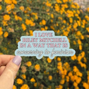May include: A light blue sticker with the text "I LOVE RILEY MITCHELL IN A WAY THAT IS concerning to feminism" in pink and white. The sticker is held in front of a background of yellow flowers.