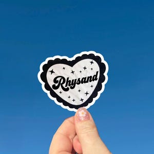 May include: A heart-shaped sticker with a scalloped white border and a black scalloped edge. The centre is white with the black text Rhysand in a cursive font, surrounded by small black four-pointed star icons scattered across the surface.
