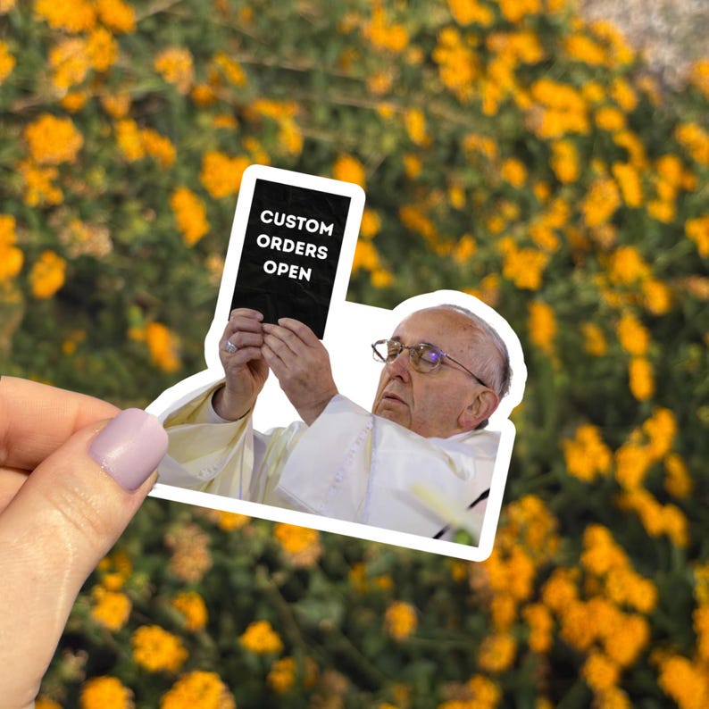 Pu&ograve; includere: Un adesivo raffigurante Papa Francesco che tiene in mano un cartello con la scritta "CUSTOM ORDERS OPEN" in bianco su sfondo nero. L'adesivo &egrave; posto su uno sfondo di fiori gialli.