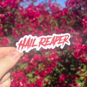 May include: A white sticker with the words "HAIL REAPER" in red, stylized lettering. The sticker is held in front of a background of vibrant pink flowers.