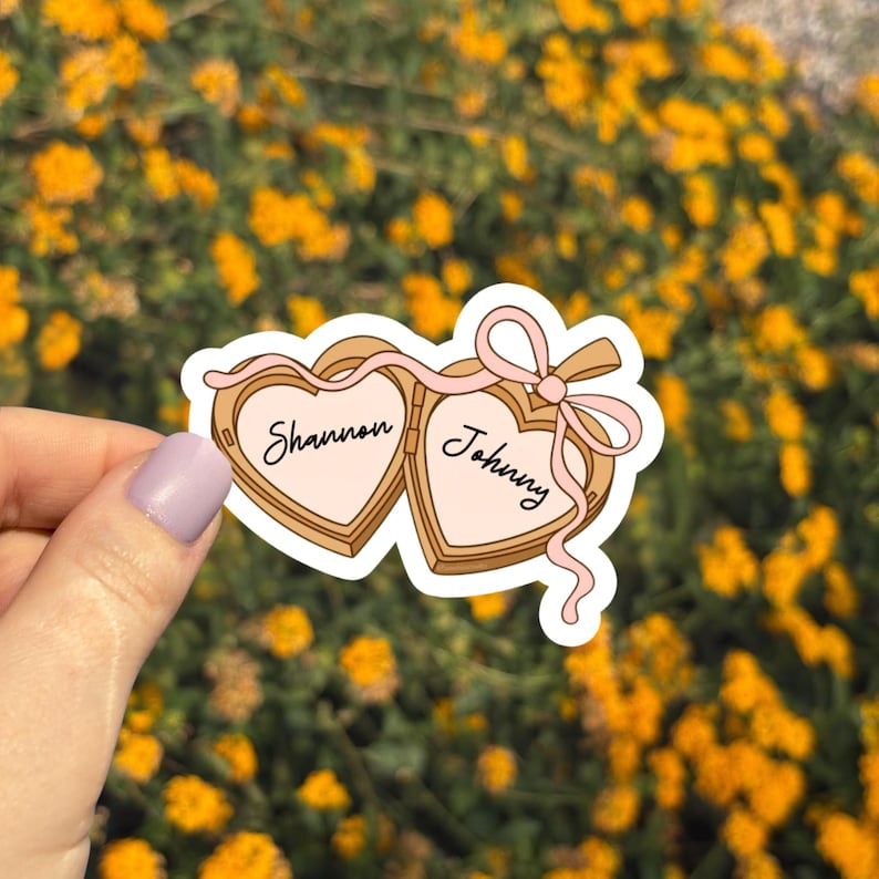May include: A sticker featuring two heart-shaped lockets, each inscribed with a name. The lockets are connected by a pink ribbon and bow. The names "Shannon" and "Johnny" are written in elegant script.