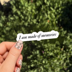 May include: A white die-cut sticker with a wavy edge featuring the black text I am made of memories in a cursive font. The sticker is held against a blurred green foliage background, showcasing a clean design for personal expression.