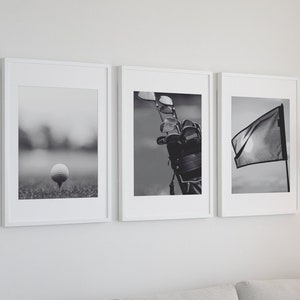 May include: Three black and white framed photographs of golf-related imagery. The first image shows a golf ball on a tee on a golf course. The second image shows a golf bag with golf clubs sticking out of it. The third image shows a golf flag waving in the wind.