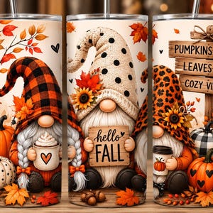 May include: Three cylindrical tumblers with metal straws featuring autumn-themed illustrations of gnomes, pumpkins, sunflowers, and fall leaves. The designs include text that reads hello FALL and PUMPKINS LEAVES COZY VIBES in a warm color palette.