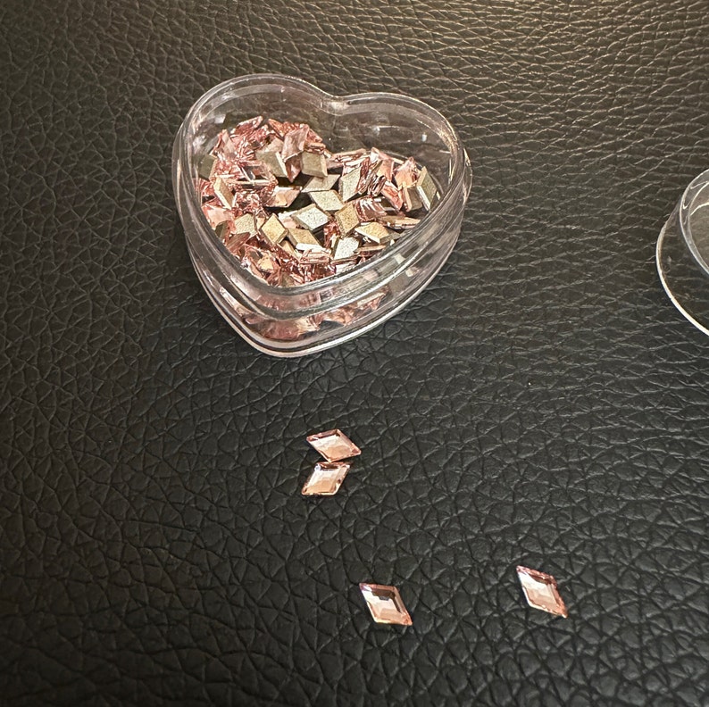 May include: A heart-shaped plastic container filled with small, rose gold-colored rhinestones. A few rhinestones are scattered on a black surface.