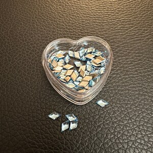 May include: A heart-shaped container filled with light blue diamond-shaped rhinestones. Some rhinestones are scattered on a brown leather surface.