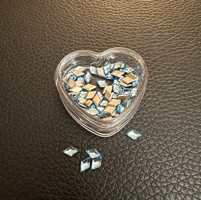 May include: A heart-shaped container filled with light blue diamond-shaped rhinestones. Some rhinestones are scattered on a brown leather surface.