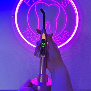 Könnte beinhalten: Ein pinkfarbenes Neon-Schild mit dem Text "Fairy's Tooth Gems" und einer Zahngrafik. Eine Person hält eine goldene und schwarze Dental-Härtungslampe mit einem digitalen Display.