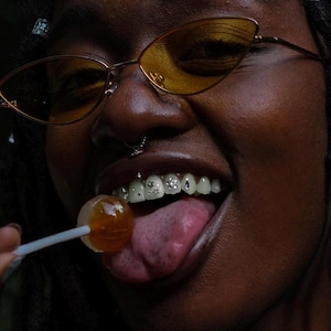 Könnte beinhalten: Nahaufnahme einer Person mit dunkler Haut, die eine goldgerahmte Sonnenbrille mit gelben Gläsern trägt. Die Person lächelt und zeigt einen Lolli in ihrem Mund und Zähne mit dekorativen Edelsteinen.