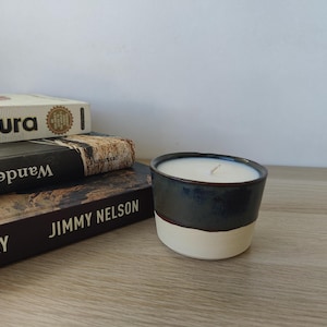 Op de afbeelding: Een handgemaakte kaars in een donkerblauwe en witte keramische houder. De kaars staat op een houten oppervlak naast een stapel boeken. De kaars heeft witte was en een enkele lont.