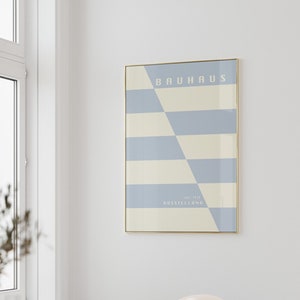 Könnte beinhalten: Gerahmtes Bauhaus-Poster mit geometrischem Design in Hellblau und Creme. Der Text "BAUHAUS" ist zu sehen. Es hängt an einer weißen Wand über einem Holztisch und einem transparenten Acrylstuhl.