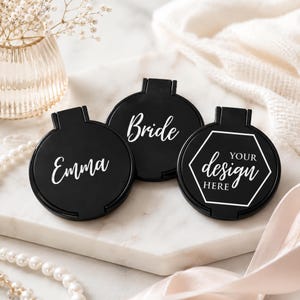 May include: Three round black compact mirrors with white text. One mirror features the name Emma, another displays the word Bride, and the third shows a white hexagonal outline with the text YOUR DESIGN HERE printed in white lettering.