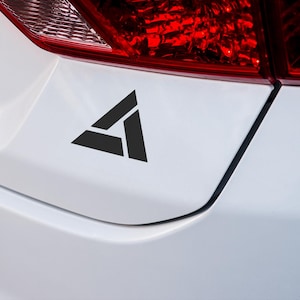 May include: A black triangle sticker with a geometric design, applied to the back of a white car.