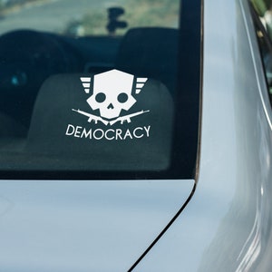 May include: A white sticker on the back window of a silver car. The sticker features a skull with wings and crossed rifles, with the word "DEMOCRACY" below it.