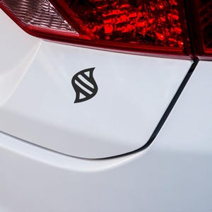 May include: Black vinyl car decal featuring a stylized leaf design. The decal is affixed to the white body of a vehicle, near the taillight. The taillight is red. The decal is a simple, yet elegant design.