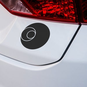 May include: A black and white circle sticker with a concentric circle design, applied to the back bumper of a white car.