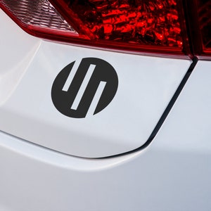 May include: A black and white circle sticker with three vertical lines inside. The sticker is on the back of a white car.