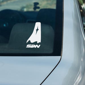 May include: White vinyl car decal featuring a stylized cape design with a star and the letters SDN. The decal is affixed to the rear window of a silver car, with the interior visible in the background.