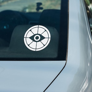 May include: A white vinyl car decal featuring a stylized eye design within a circular frame. The decal is affixed to the rear window of a silver car. The design has a geometric pattern and is likely for vehicle customization.