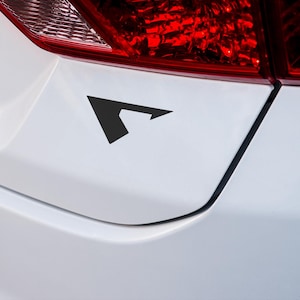 May include: A black arrow-shaped decal is affixed to the white surface of a vehicle. The decal is positioned near the red taillight, adding a graphic element to the car's exterior.