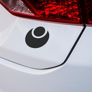 May include: A black and white sticker of a stylized circle with a smaller circle inside, placed on the back of a white car.