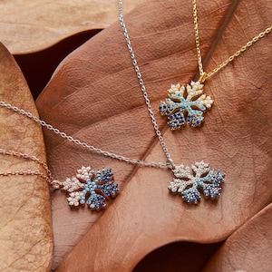 Puede incluir: Tres collares de plata con forma de copo de nieve con detalles de cristal azul y blanco sobre un fondo marrón. Los collares están sobre una hoja marrón con el texto "Selin Silver" en la esquina inferior derecha.