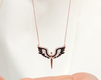 May include: A rose gold necklace featuring a black and white angel pendant with clear crystal accents. The pendant depicts a stylised angel with wings and a pointed base. The necklace is on a chain with a delicate design.