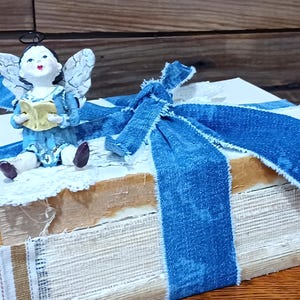 May include: A decorative stack of books, painted white, with a blue denim ribbon tied around them. A small angel figurine in blue and white sits on top, holding a book. The books are trimmed with lace and tan paper.