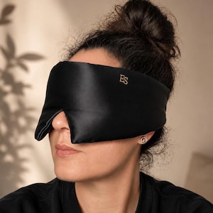 May include: A black sleep mask with a smooth, satin-like finish and a contoured shape. The letters ES are embroidered in gold thread on the upper right side of the mask. The mask covers the eye area and features a nose bridge cutout.