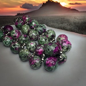 May include: A strand of green and pink gemstone beads with silver accents. The beads are round and have a speckled pattern.