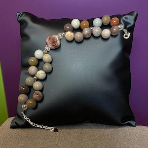 Puede incluir: Una pulsera de cuentas con una variedad de cuentas en tonos tierra, incluyendo blanco, marrón y gris. La pulsera presenta una cuenta en forma de rosa y un cierre de cadena plateada. La pulsera se exhibe sobre un cojín negro sobre un fondo púrpura.
