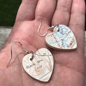 May include: A pair of heart-shaped earrings made from maps. The left earring features a map with the text "Black Mtn" and contour lines. The right earring features a map with the text "Hill Alder" and a blue river.