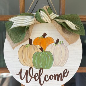 May include: A round wooden door hanger with a burlap and green ribbon bow. The door hanger is decorated with painted pumpkins in shades of orange, green, and white. The words "Welcome" are painted in brown on the door hanger.