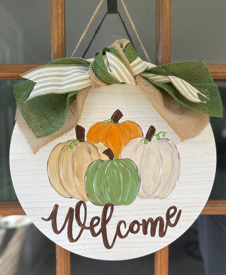 May include: A round wooden door hanger with a burlap and green ribbon bow. The door hanger is decorated with painted pumpkins in shades of orange, green, and white. The words "Welcome" are painted in brown on the door hanger.