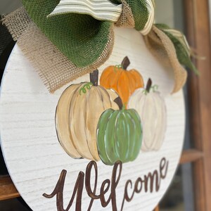 May include: A round wooden door hanger with a white background and a brown welcome sign. The door hanger is decorated with four painted pumpkins in shades of orange, white, green, and beige. The pumpkins are surrounded by a burlap bow with a green and white striped ribbon.